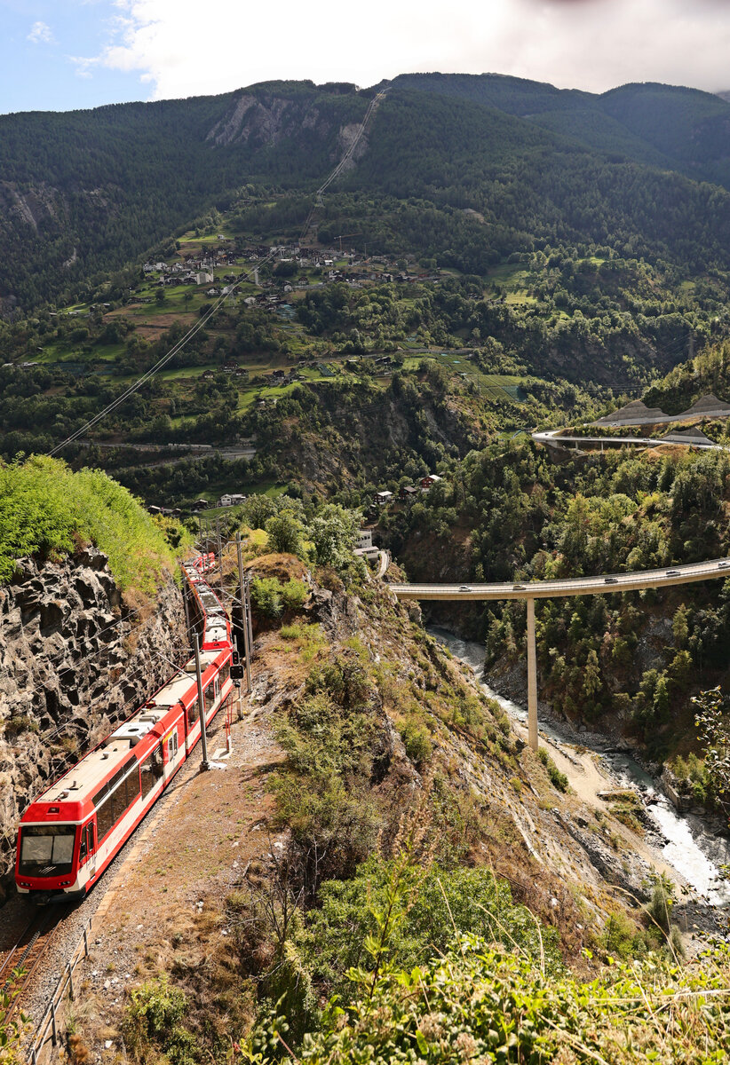 Abstieg eines MGB-Zuges von Zermatt her nach Stalden. Der lange Zug ist zusammengesetzt aus ABDeh4/10 2011 + ABDeh4/8 2025 + Doppelsteuerwagen ABt 2131. Blick auf die hohe Brücke der Umfahrungsstrasse Richtung Täsch/Saas Fee. 30.August 2025  