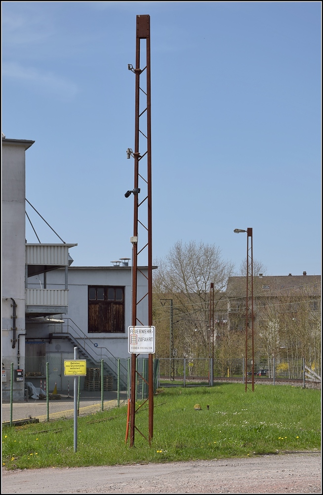 Anschlussverwendung der Oberleitungsmasten der badischen Staatsbahn in Lörrach-Brombach an einem ehemaligen Werksgleis. Kamerainstallation und Lampenmast. April 2013

Die ersten ernstzunehmenden Elektrifierzierungen mit Einphasenwechselstrom wurden anfangs mit 15 kV und 15 Hz durchgeführt. Durch einen Beschluss der badischen, bayrischen und hessisch-preußischen Staatsbahnen im Jahr 1912 wurden 16 2/3 Hz festgelegt. Die BLS schloss sich dem an und stellte ihre bereits elektrifizierte Strecke gleich darauf um, die RhB verwendete ebenso 16 2/3 aber eine reduzierte Spannung von 11 kV, die SBB hingegen begann erst ab 1919 mit Elektrifizierungen. In Deutschland waren Wiesen- und Wehratalbahn das größte elektrifizierte Netz ab 1913, immerhin bis 1936 mit 15 Hz. Der 1. Weltkrieg bremste erst einmal weitere Elektrifizierungen bis 1922 aus. Somit dürften diese Masten 105 Jahre alt sein. Kurz vor Zell im Wiesental sind sogar noch einige dieser Masten in Betrieb. Im Bild der vierte Mast ist aus der Reichsbahnzeit.