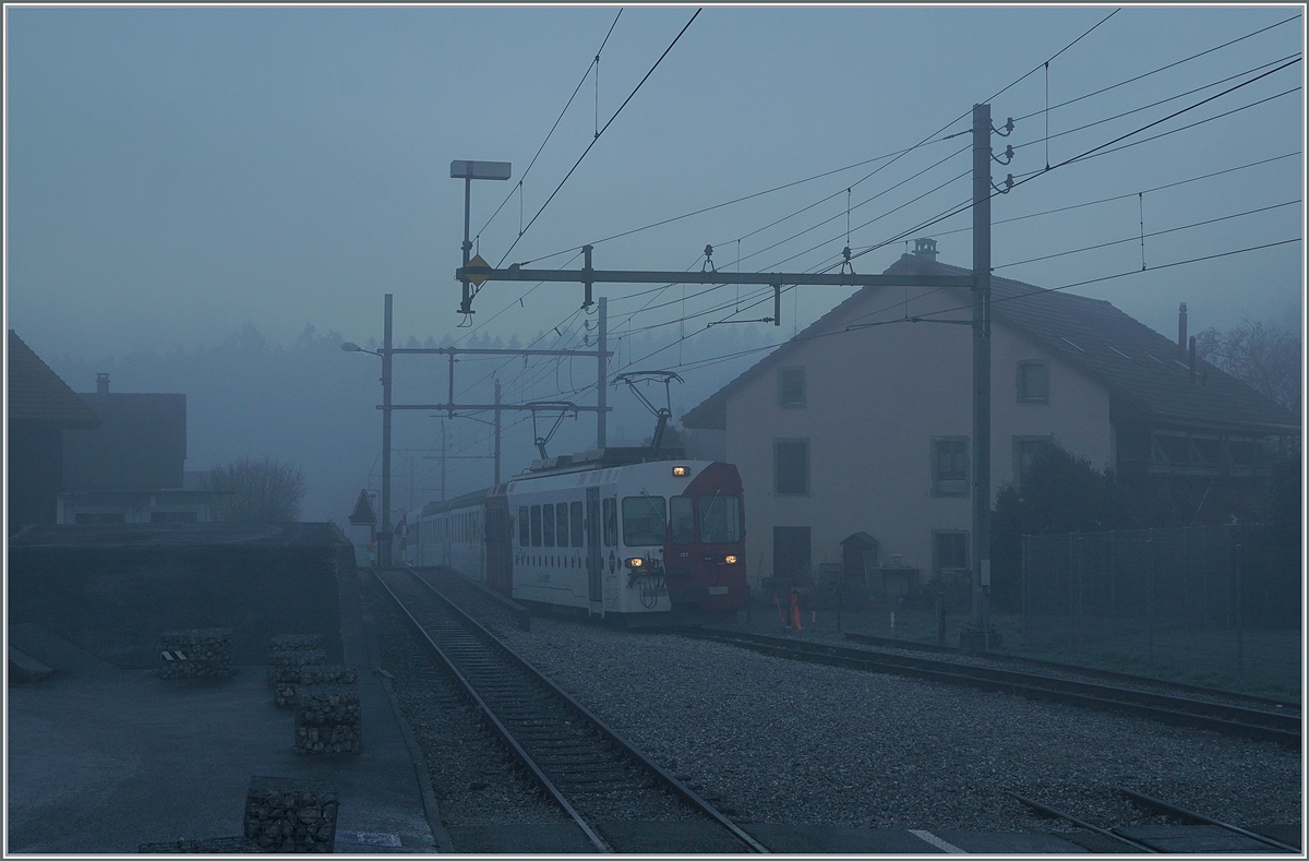 Bei dichtem Nebel und noch in der Morgendämmerung erreicht der TPF Be 4/4 121 mit dem B 207 (ex MOB), B209 (ex MOB) und dem ABt 221 den Bahnhof von Broc Village. Der Zug ist als RER 60 14954 von Bulle nach Broc Farbrique unterwegs. 

26. Nov. 2020