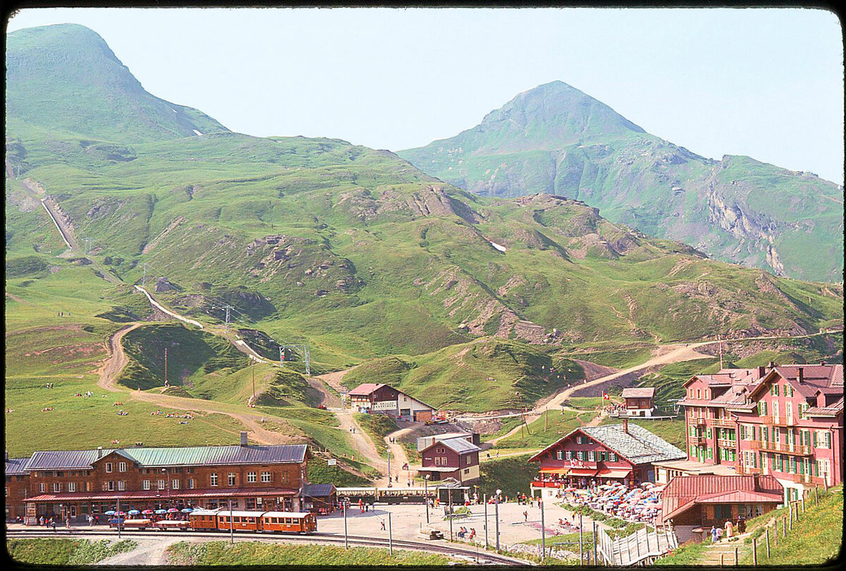 Blick auf die Kleine Scheidegg mit alten Zügen: Bei der Jungfraubahn ein Zug mit Lok 11, bei der Wengernalpbahn ein Zug Richtung Grindelwald mit Lok 51. 5.August 1975 