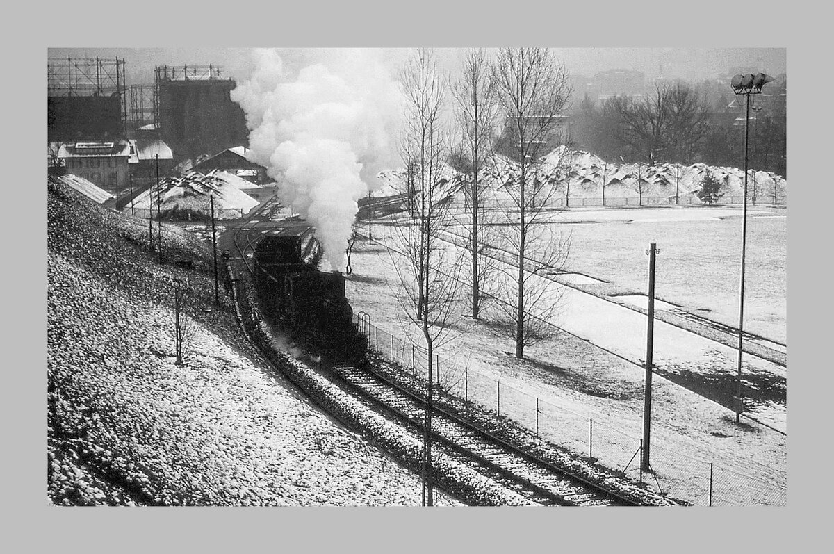 Die Gaswerkbahn Bern: Die Dampflok verlässt das ehemalige Gaswerk-Areal am Aareufer, um die 35 Promille-Steigung nach Wabern hinauf in Angriff zu nehmen. Die Gaskessel dienen heute als Jugend-Treff, der Geruch von Kohle und Gas (?) ist nur noch eine ferne Erinnerung, die sich jedoch unangenehm ins Gedächtnis gebrannt hat. Dezember 1964  