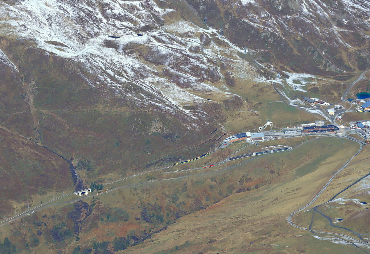 Ein scharfer Blick vom Jungfraujoch hinunter. Auf der Kleinen Scheidegg sind zu sehen: Ein WAB-Zug mit zwei Panorama Triebwagen, der in Richtung Lauterbrunnen wegfährt, ein JB-Dienstzug vor dem unteren Depot, mit einem der beiden noch verbliebenen Vierachs-Triebwagen 204 oder 206 + Werkstattwagen X 55, ein alter WAB-Vierachszug (Triebwagen 114 + Steuerwagen Bt 266), wie er sonst auf der WAB nicht mehr zu sehen ist. Dazu kommt noch ein Blick auf die Spitzkehre der WAB im Berg drin, der den Uebergang der Zuege von der Lauterbrunnen-Seite zur Grindelwald-Seite (bzw. umgekehrt) ermöglicht. 14.Oktober 2021  