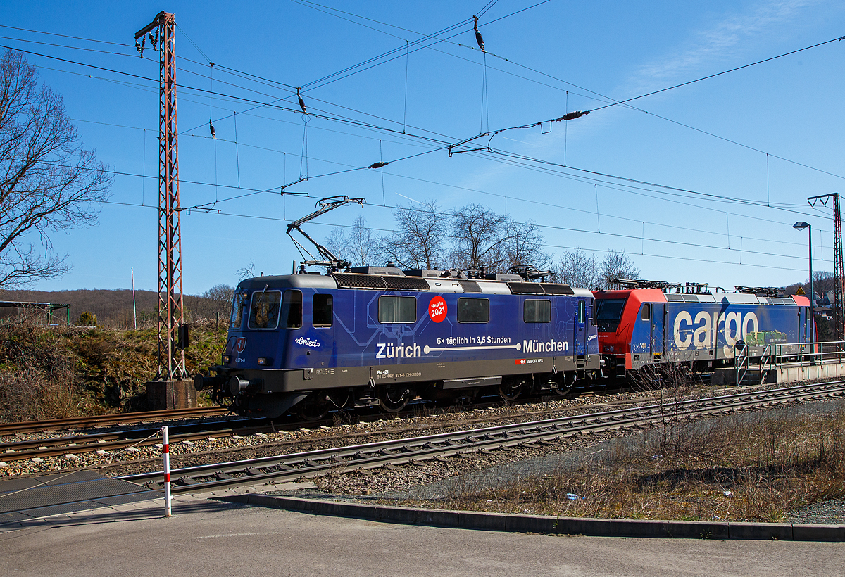 Ja, „Grüezi“ oder „Servus“.....
Die SBB Cargo Re 421 371-6 (91 85 4421 371-6 CH-SBBC), ex Re 4/4 II 11371, mit Werbung Zürich – München 6 x täglich in 3,5 Stunden, fährt am 30.03.2021 mit der SBB Cargo Re 484 016-1(91 85 4484 016-1 CH-SBBC), ex E 484.016 SR, einer TRAXX F140 MS am Haken durch Rudersdorf (Kr. Siegen), an der Dillstrecke (KBS 445), in Richtung Siegen.

Eine SBB Cargo Re 484 wie die 016-1 kann man eigentlich Deutschland nur mit viel Glück sehen, denn sie haben nur die Zulassungen für die Schweiz und Italien. Sie haben keinen Stromabnehmer mit breiterer Wippe gemäß deutscher Vorschrift, auch die die deutsche Zugsicherung Indusi fehlt ihnen. Der Lz war auf dem weg nach Krefeld.


Lebensläufe:
Die Re 421 371-6 wurde 1983 von der SLM Winterthur (Elektrik BBC/ SAAS) unter der Fabriknummer 5235 gebaut und als Re 4/4 II 11371 an die SBB geliefert. Im Jahr 2002 erfolgte der Umbau zur Re 421 für die SBB Cargo (Einsatz auch in Deutschland). Die zugelassene Höchstgeschwindigkeit für die Schweiz beträgt 140 km/h und für Deutschland 120 km/h.

Die Re 484 016-1 eine Bombardier TRAXX F140 MS wurde 2004 von Bombardier in Kassel unter der Fabriknummer 34009 gebaut und an die SBB Cargo AG, als E 484.016 SR geliefert. 
