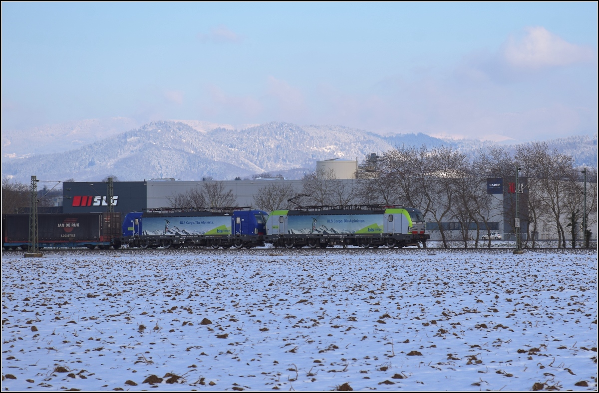 Zwischen Belchensystem und Blauendreieck. 

Die neue (vorübergehende) Fotostelle erlaubt schöne Fernblicke hinter dem Zug. Hier zieht Re 475 409 einen Zug Richtung Basel mit der 193 496 der BLS im Schlepptau. Buggingen, Februar 2021.