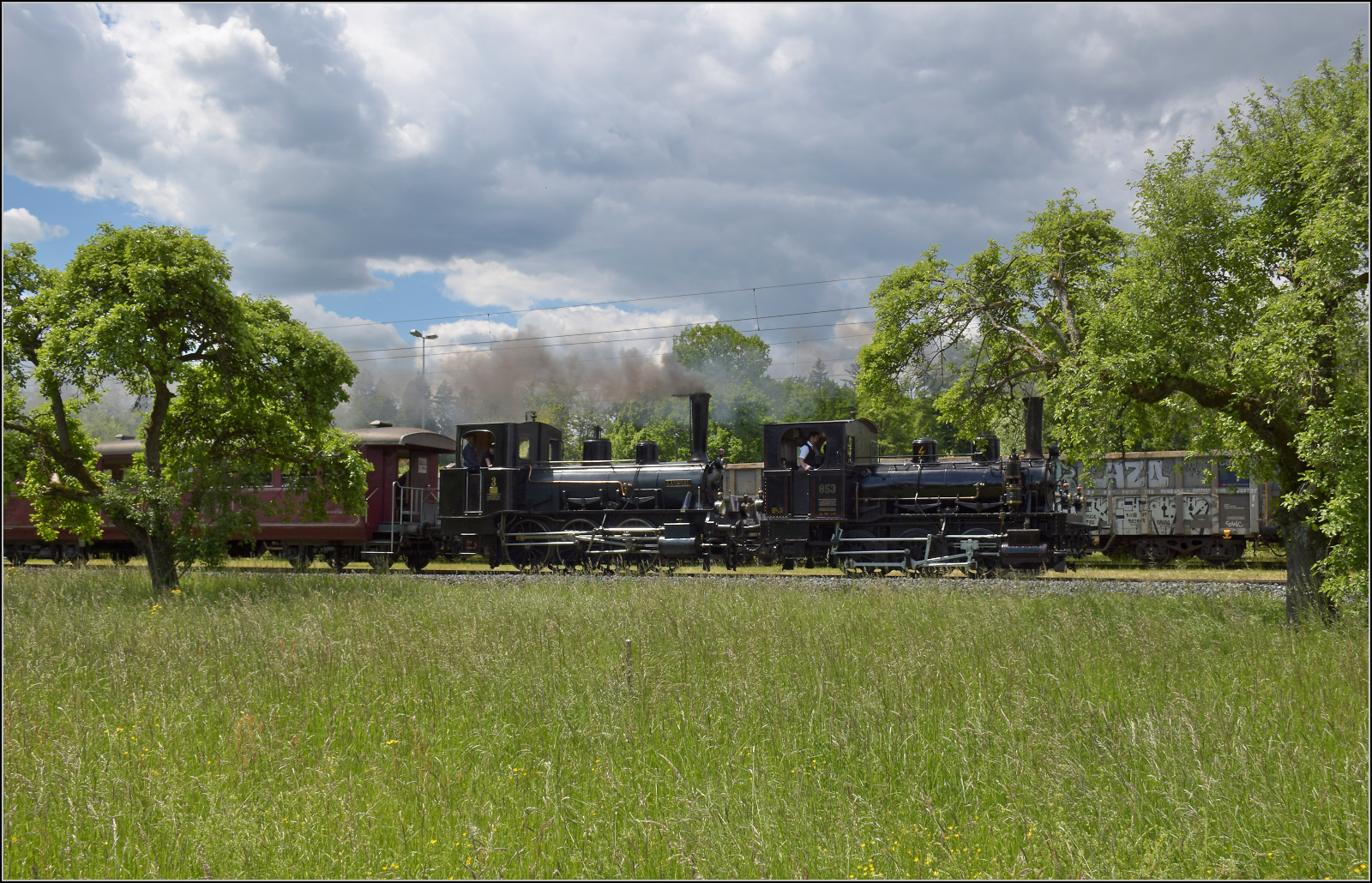 150 Jahre Emmentalbahn. 

E 3/3 853 der Jura-Simplon und Ed 3/3 3 'Langnau' der Emmentalbahn fahren mit ihrem Sonderzug durch einen Obstgarten in den Schwerindustriestandort Gerlafingen ein. Mai 2025.
