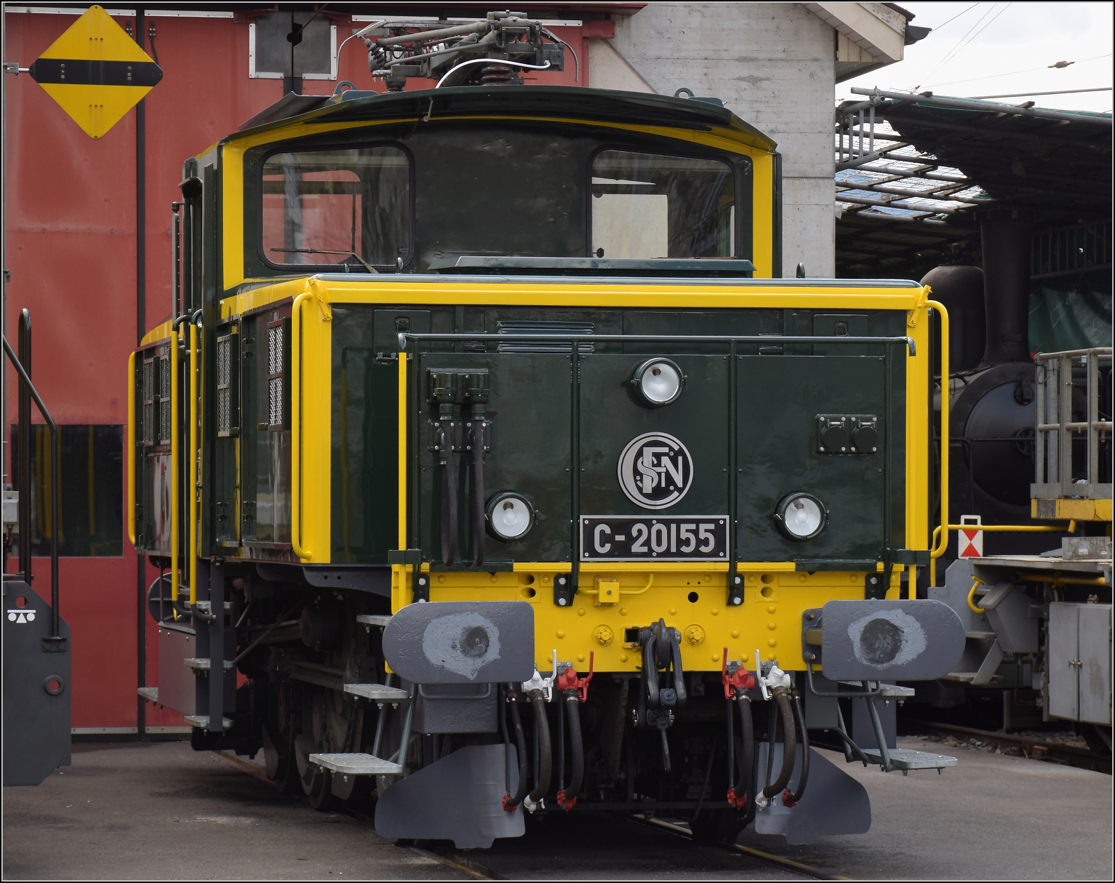 Etwas versteckt vor der Werkstatt in Balsthal steht die wunderbar in den Originalzustand zur�ckversetzte Zweispannungsrangierlok Ee 3/3 16515. Die letzte erhaltene Rangierlok dieser Baureihe war zu Beginn ihrer Karriere von den SNCF trotz sehr niedriger H�chstgeschwindigkeit im Streckendienst nach Basel eingesetzt. Weil sie doch im Rangierdienst besser geeignet ist, kam sie ein Jahrzehnt sp�ter zu den SBB als Erg�nzung in den Rangierdienst. Ende letzten Jahres bekam sie ihr altes Farbkleid und die Bezeichnung C 20155 zur�ck. Nicht ganz unumstritten, aber meiner Meinung nach eine sch�ne Idee und wunderbar anzusehen. 

Dahinter steckt noch eine der kleinen Dampfloks der OeBB historic ihre Rauchkammert�r an die frische Luft. Balsthal, April 2025. 